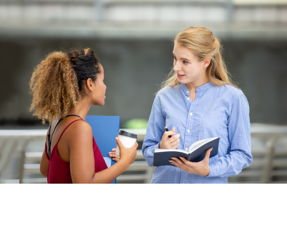 Zwei Frauen im Gespräch miteinander. Die eine hält ein aufgeschlagenes Buch in der Hand, die andere ein Schreibheft und einen Kaffeebecher